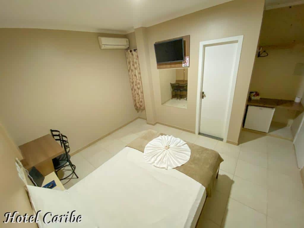 Quarto do Hotel Caribe. Do lado direito uma prateleira, e um frigobar. No meio uma cama de casal e de frente a porta do banheiro, televisão e espelho. No lado esquerdo uma mesa de trabalho e um ar-condicionado. Foto para ilustrar post sobre hotéis em Porto Velho.