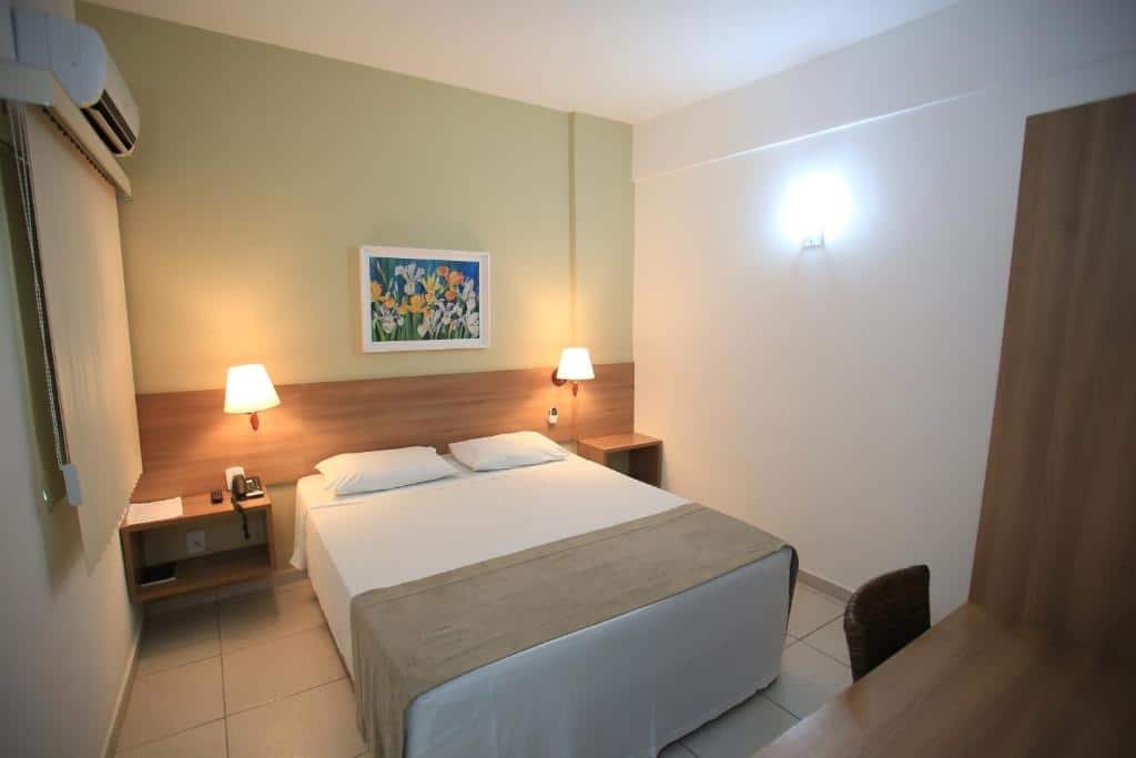 Quarto do Hotel Ecos Classic. Uma mesa de trabalho do lado direito, do lado esquerdo uma cama de casal com uma prateleira e um abajur de cada lado. No lado direito a janela com cortina e ar-condicionado.