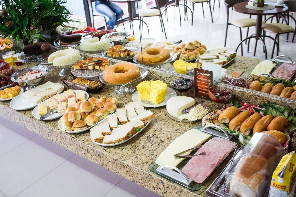 Balcão do café da manhã do Hotel Nativo. Uma mesa com frios, pães e bolos. Em cima um espelho refletindo as mesas e cadeiras.