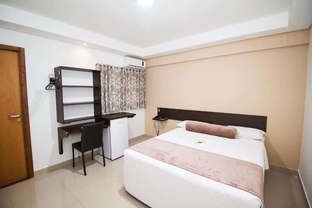 Quarto do Hotel Nativo. Do lado direito uma cama de casal, do lado esquerdo a porta do quarto, uma mesa de trabalho com uma prateleira, um frigobar em cima da janela um ar-condicionado.