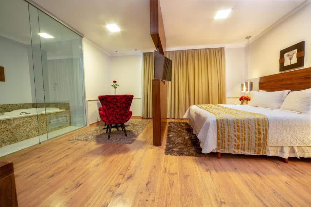 Quarto do Hotel Porto Madeira. Do lado direito uma cama de casal, no meio um painel com uma televisão. Do lado esquerdo uma banheira de hidromassagem e uma poltrona.