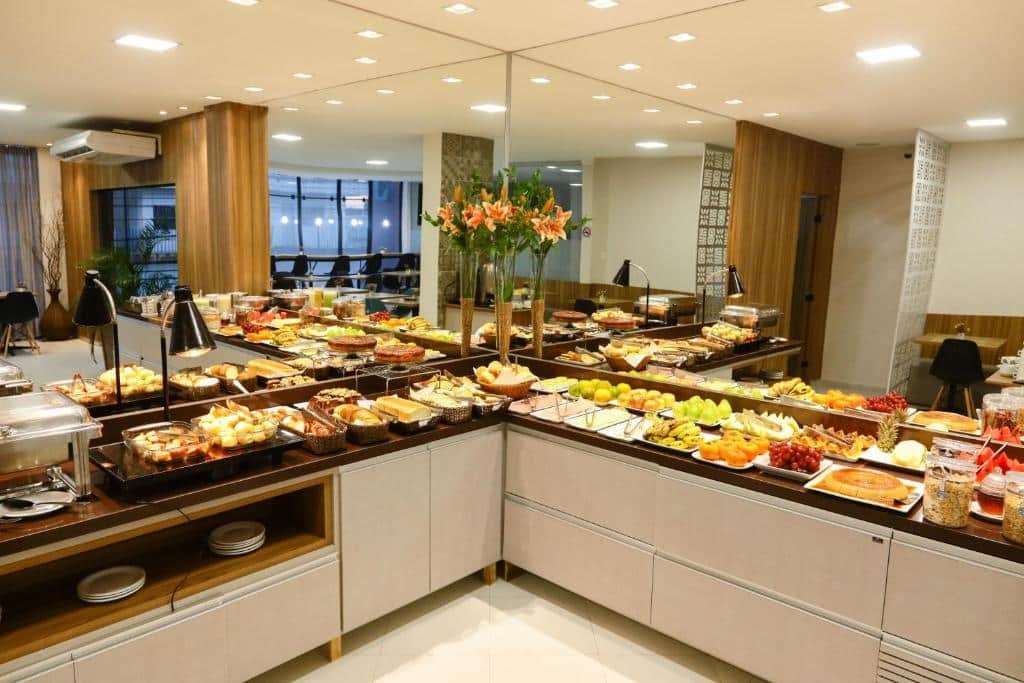 Balcão do café da manhã do Hotel Porto Madeira. Uma mesa cheia de frutas, frios, pães e doces, em cima uma parede espelhada, em baixo um armário.