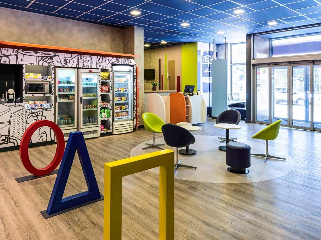 Recepção do hotel com cadeira coloridas, um círculos, triângulo e quadrado em vermelho, azul e amarelo, geladeiras com coisas de beber e uma porta de vidro.