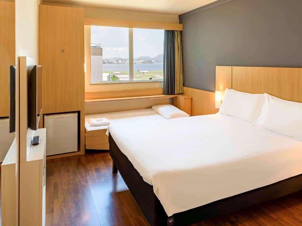 Quarto com uma cama de casal e uma para criança, frigobar, janela de vidro, tv na parede e chão de madeira, ilustrando post hotéis Ibis no Rio de Janeiro.