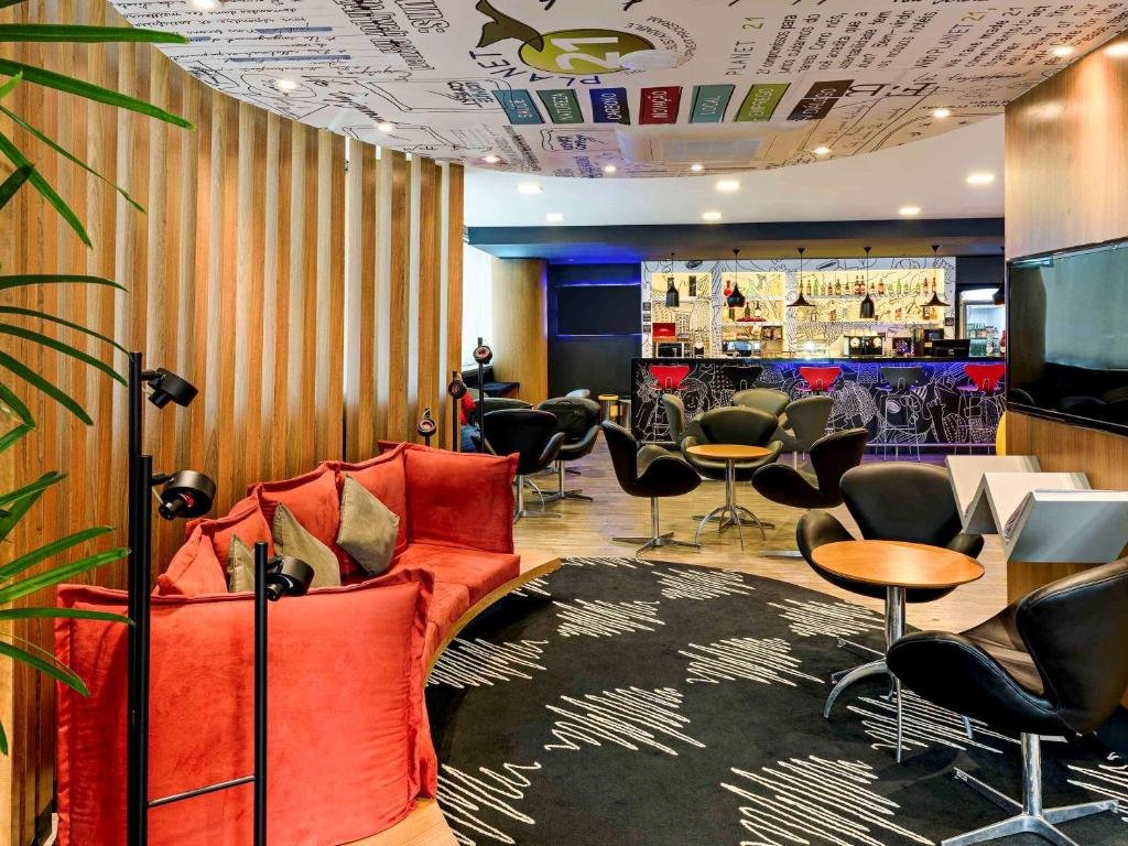 Espaço interno do hotel com sofá vermelho, algumas mesas com cadeiras pretas, tapete preto com detalhes em branco, tv na parede, detalhes em madeira e teto colorido.