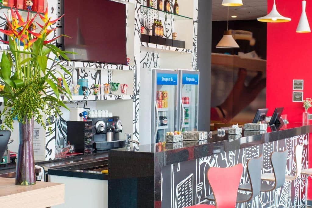 Balcão na cor preta com cadeiras vermelhas, pretas e branca e na frente uma parede toda desenhada em preto e branco, com duas geladeiras, tv e um vaso de planta.