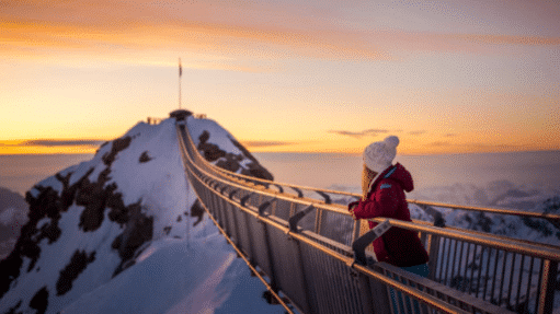 Uma pessoa com roupas de inverno em Glacier que está em uma ponte nevada na montanha ao pôr do sol, olhando para o pico distante.