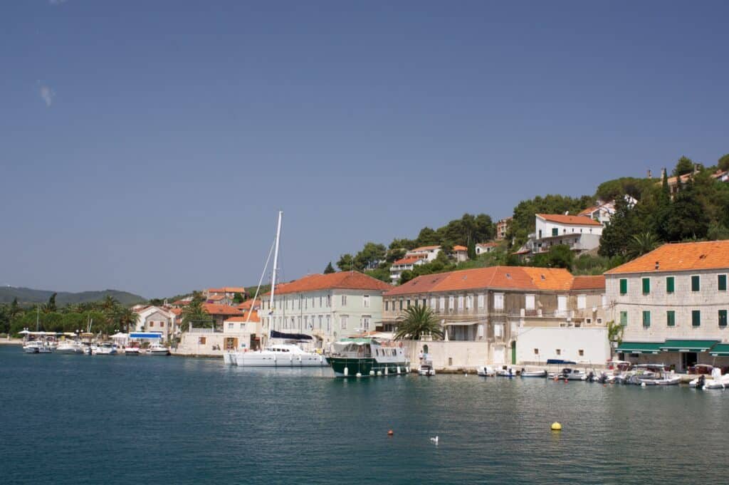 Uma vila costeira em Jelsa com barcos atracados à beira-mar, cercada por prédios com telhados vermelhos sob um céu azul claro.
