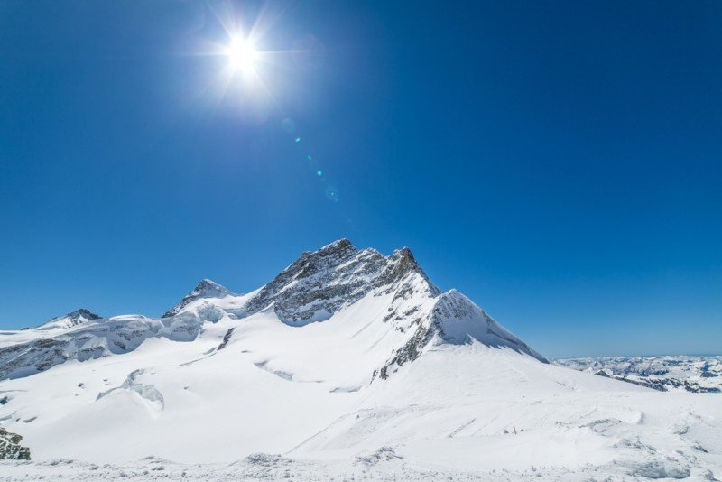 Montanha Jungfrau, coberta pela neve. O céu é azul e o sol brilha no topo.