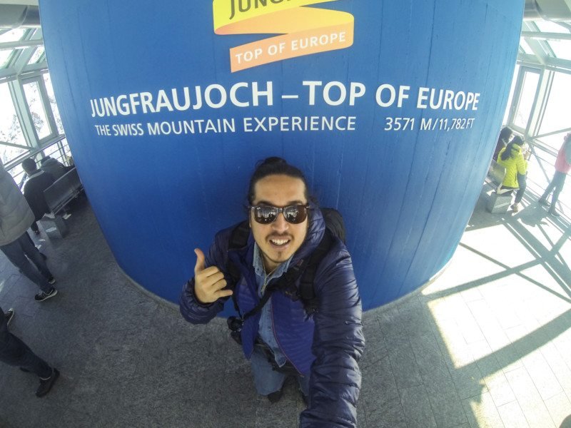 Foto de um homem de óculos escuros e roupas de inverno fazendo sinal de "hang loose" em frente a uma parede azul da estação de Jungfraujoch.