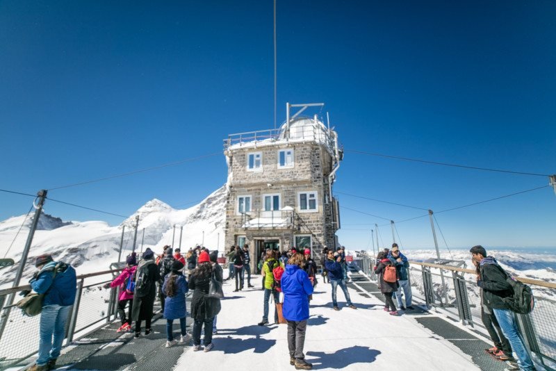 Muitas pessoas reunidas em frente ao Observatório Sphinx, em Jungfrau. Está de dia e o céu é bem azul.