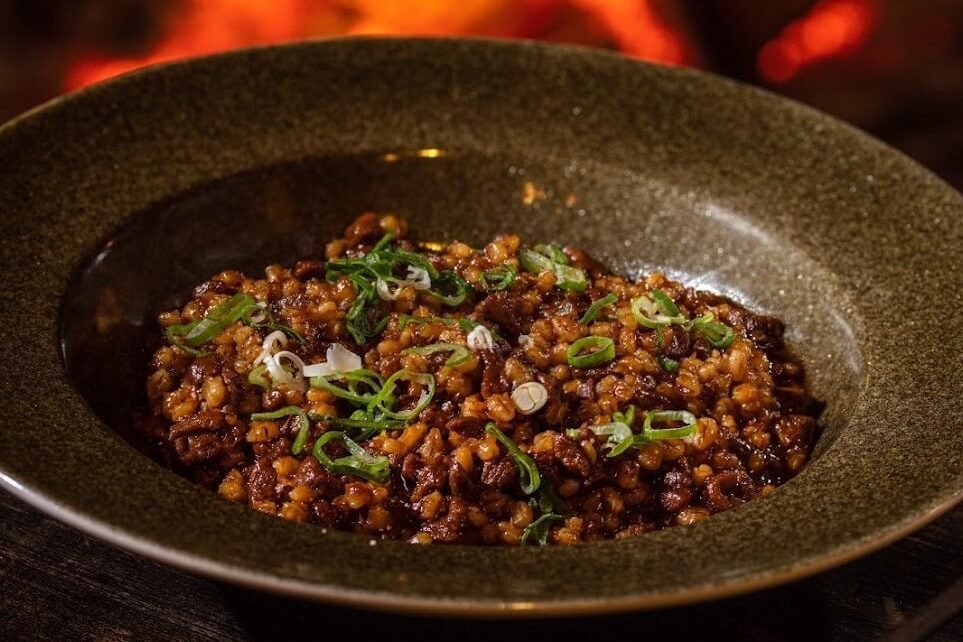 Uma tigela de um prato não identificado, coberta com cebolinha verde é colocada sobre uma mesa de madeira escura com uma fogueira acesa ao fundo no Restaurant Lička Kuća.