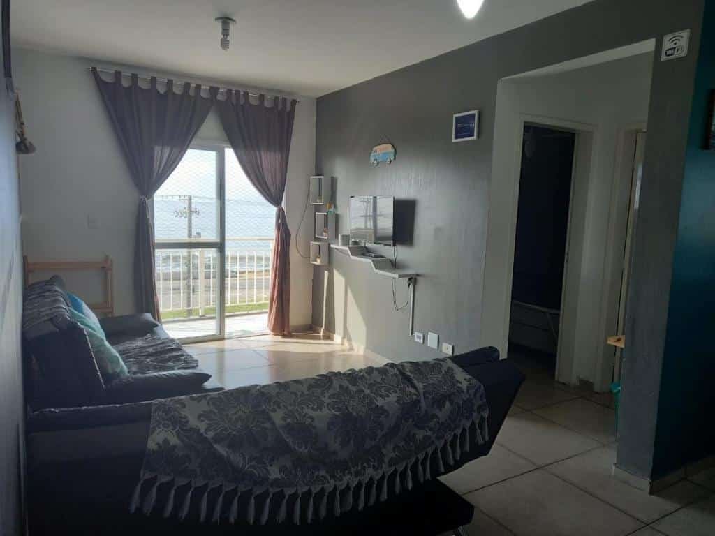 Sala de estar da Linda Vista e Som das Ondas. Dois sofás com almofadas e mantas encaram uma TV na parede ao lado direito, que tem portas para os quartos. Ao fundo há uma porta de vidro com cortinas que leva à varanda com vista do mar.