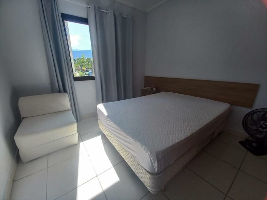 Quarto do Lindo apartamento com vista para o mar em Caraguá!, um dos airbnb em Caraguatatuba. Uma cama de casal está encostada ao lado direito do quarto, com uma mesinha de cabeceira ao lado esquerdo e um ventilador ao lado direito. Na parede lateral da cama fica uma janela com cortinas, e uma poltrona está bem ao lado.