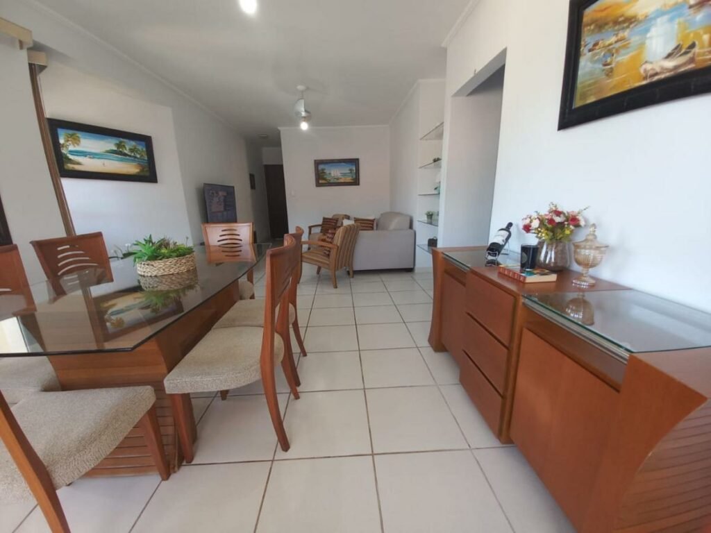 Sala de jantar do Lindo apartamento com vista para o mar em Caraguá! Ao lado esquerdo está uma mesa com seis cadeiras, e ao lado direito fica um móvel com decorações. Ao fundo é possível ver uma sala de estar com sofás e poltronas encarando uma TV.