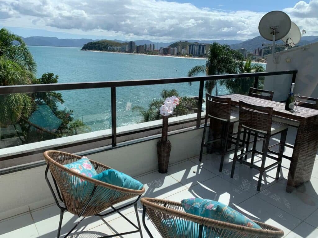 Varanda do Lindo Duplex Pé Na Areia com Praia Privativa. Duas cadeiras de jardim encaram uma mesinha com quatro cadeiras altas. Ao lado esquerdo há uma borda de vidro com vista para o mar.