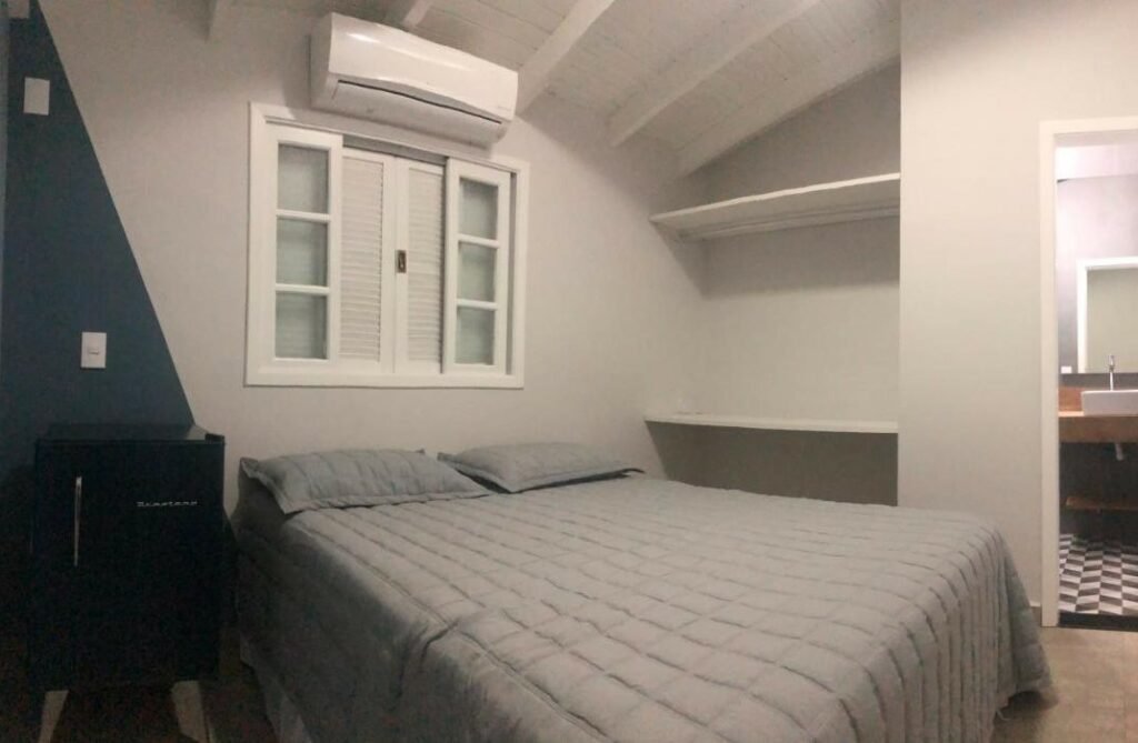 Quarto do Lindo Flat em Maresias, 100m da Praia. Uma cama de casal está no meio, abaixo de uma janela e ao lado de uma mini geladeira. Do outro lado estão duas prateleiras na parede e um espelho.