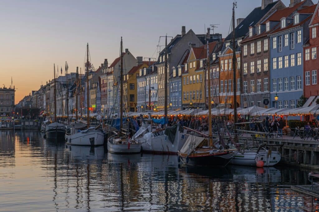 Barcos atracados ao longo de um canal em Nyhavn, Copenhague, ladeado por edifícios coloridos ao pôr do sol, com pessoas sentadas em cafés ao ar livre em primeiro plano. Esse é um dos destinos indicados no guia de mochilão pela Europa.
