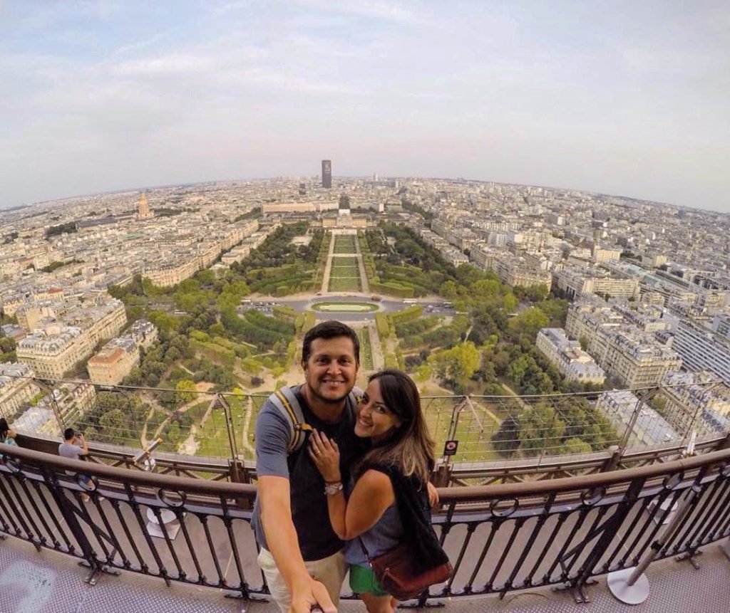 um casal abraçado na beirada da amurada, no alto da torre eiffel em Paris, sorrindo para câmera. Atrás deles é possível ver todo o jardim da cidade, além de várias ruas e prédios históricos