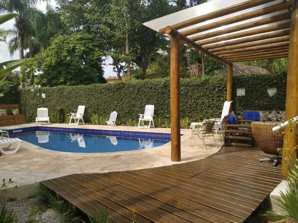 Piscina da Mares de Camburizinho rodeada por cadeiras e espreguiçadeiras brancas. Na lateral direita está uma área de deque coberta e com móveis de jardim. Representa airbnb em São Sebastião.