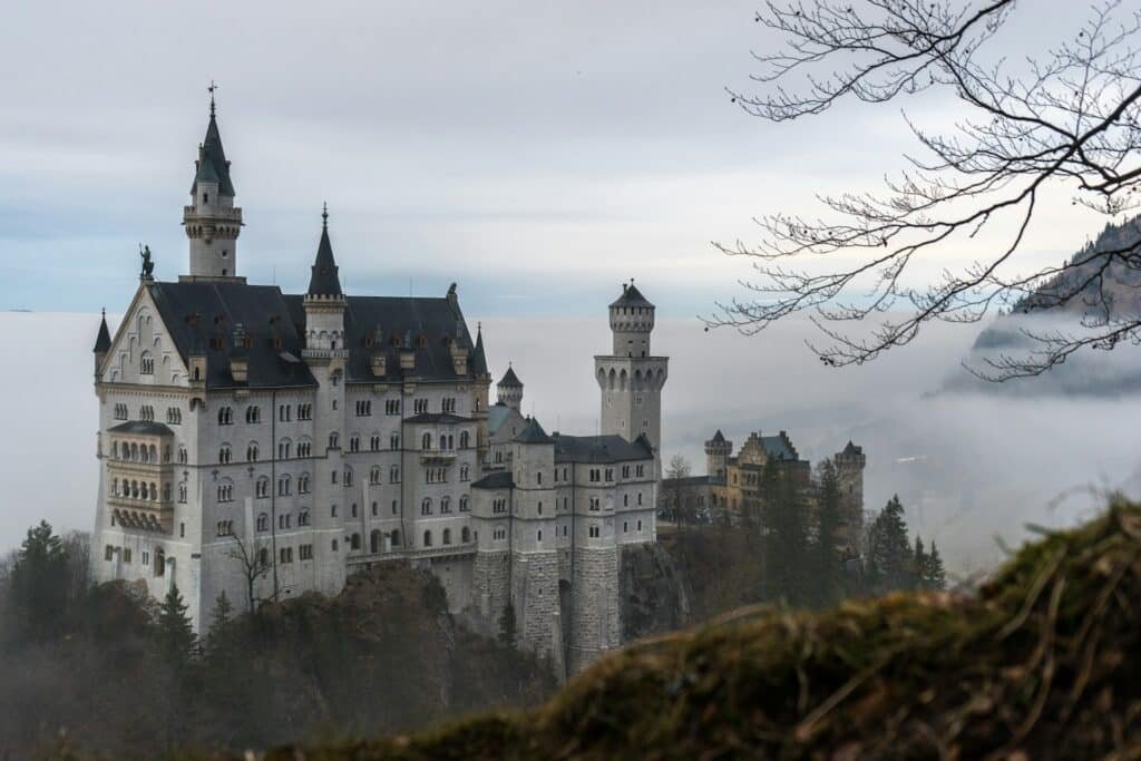 Um grande castelo histórico com torres altas fica em uma colina cercada por névoa e árvores, sob um céu nublado. Esse é o Castelo de Neuschwanstein.