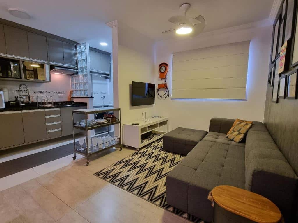 Foto de um apartamento em Novíssimo Apto em Águas de São Pedro. O ambiente é espaçoso, possui um sofá cinza largo com poltrona e um rack de TV em sua frente. A televisão está fixa na parede. Os cômodos são abertos e uma cozinha completamente equipada está atrás da parede da televisão. Possui armários, pia, micro-ondas, e outros utensílios necessários.