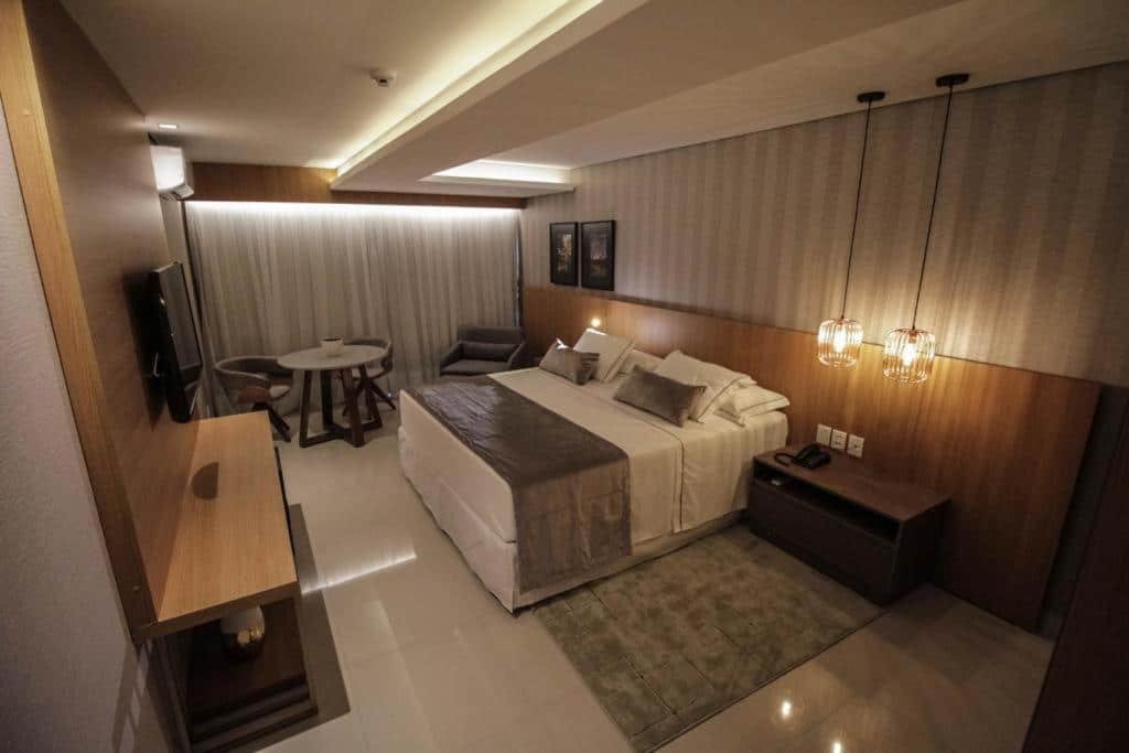 Quarto do Oscar Hotel Executive. Do lado direito uma cama de casal, com duas luminárias, uma cômoda e uma poltrona. Do lado esquerdo um painel de madeira com uma televisão, no fundo uma mesa redonda com duas cadeiras. A parede do fundo fechada com cortina.