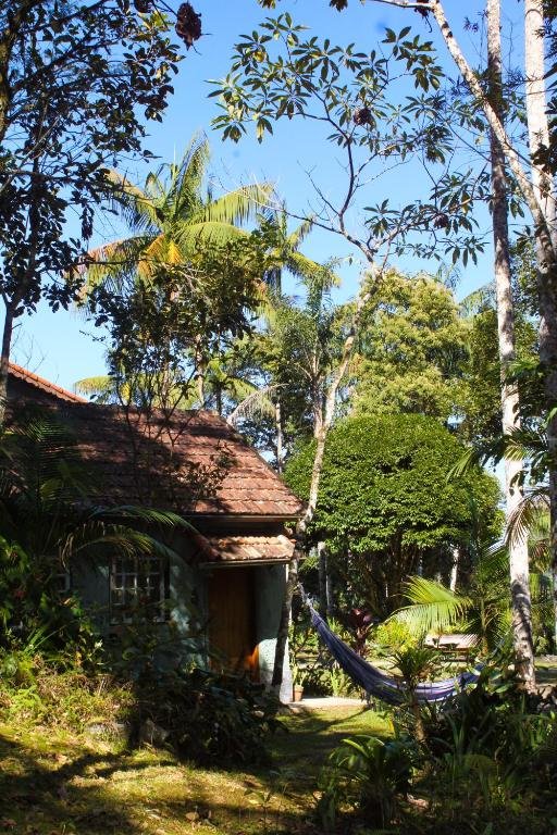 Chalé Gaia visto do lado de fora. O chalé está posicionado no lado esquerdo da imagem e no lado direito há uma rede de descanso entre duas árvores. Tanto o chalé quanto a rede estão em volta de uma área verde cheia de árvores e arbustos.