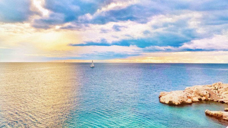 Um veleiro em um mar calmo em Ístria ao pôr do sol, com uma costa rochosa em primeiro plano e um céu nublado ao fundo. Esse é um dos destinos indicados no guia de mochilão pela Europa.