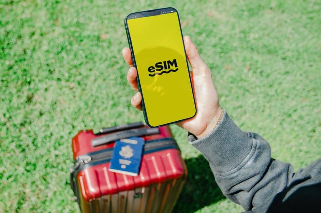 Uma pessoa segura um smartphone com a inscrição "eSIM" em uma tela amarela, acima de uma mala vermelha e um passaporte, na grama verde. A foto faz parte do post de eSIM Croácia.