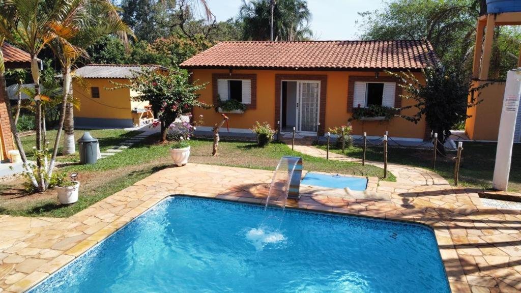 Foto da parte exterior da pousada Família Busca-Pé, em São Pedro. Vemos parte da pousada, há uma porta branca que possui um caminho de pedras até o deck da piscina. A piscina é larga e possui um chafariz.