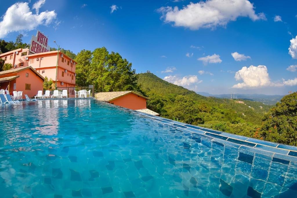 Hotel Refúgio Vista Serrana. Piscina azul de borda infinita, no lado direito a vista da serra e o céu azul claro. No fundo da imagem um pedaço do hotel laranja. Foto para ilustrar post sobre Pousadas na Serra da Cantareira