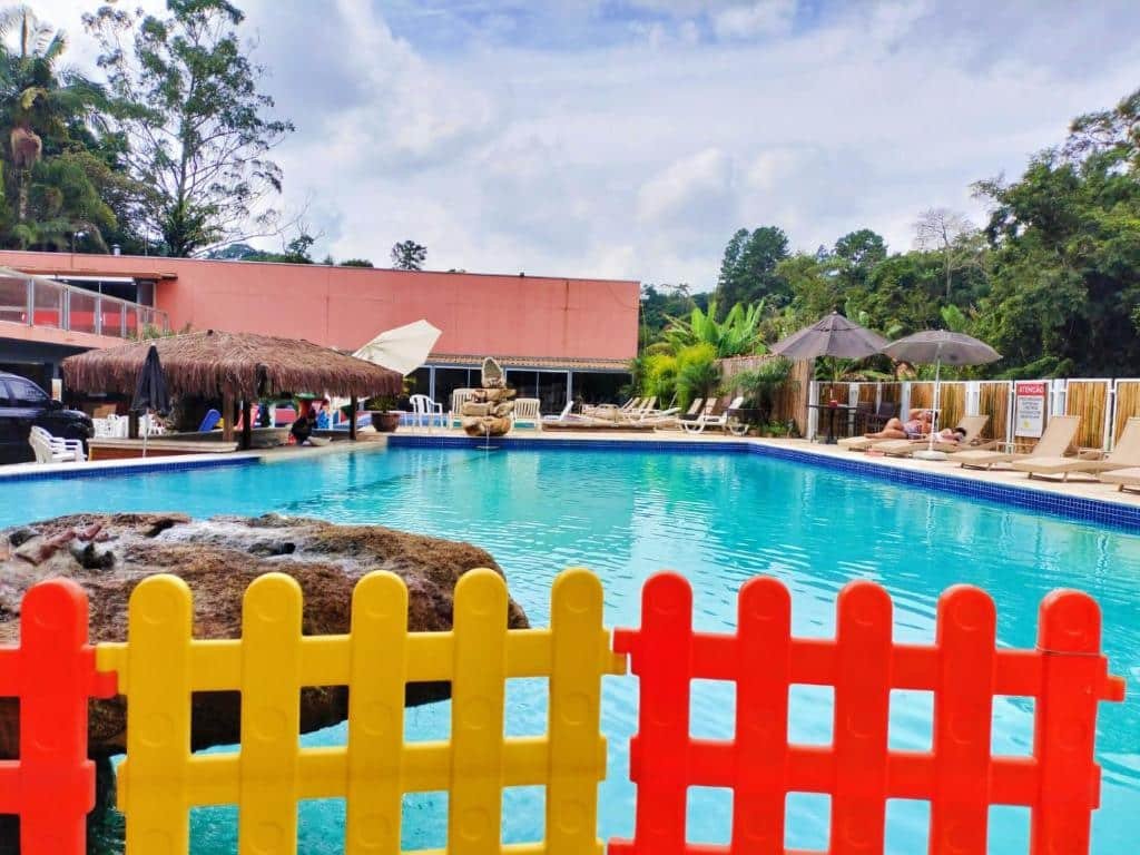 Piscina na Pousada Vale do Sol. Um cercado para as crianças vermelho e amarelo. Atrás a piscina, ao redor cadeiras de tomar sol, no canto direito um bar com acesso para a piscina. Foto para ilustrar post sobre Pousadas em Embu das Artes.