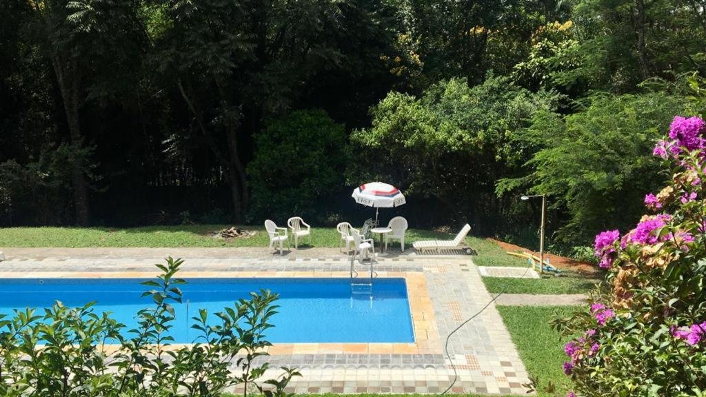 Vista da janela do quarto para a piscina na Toca da Capuava. Do lado da piscina um guarda-sol, uma mesa e cadeiras.