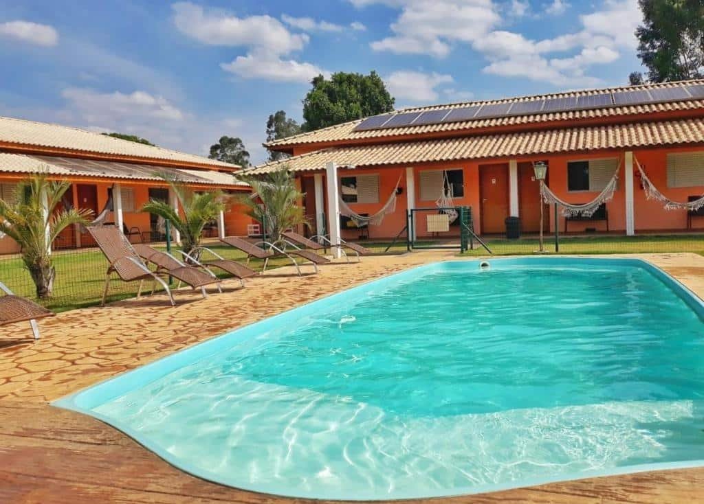 Uma piscina grande na Pousada San Pietro. Algumas espreguiçadeiras estão ao seu redor no deck. No fundo podemos ver as portas de acesso para os quartos, todas com uma rede de descanso.