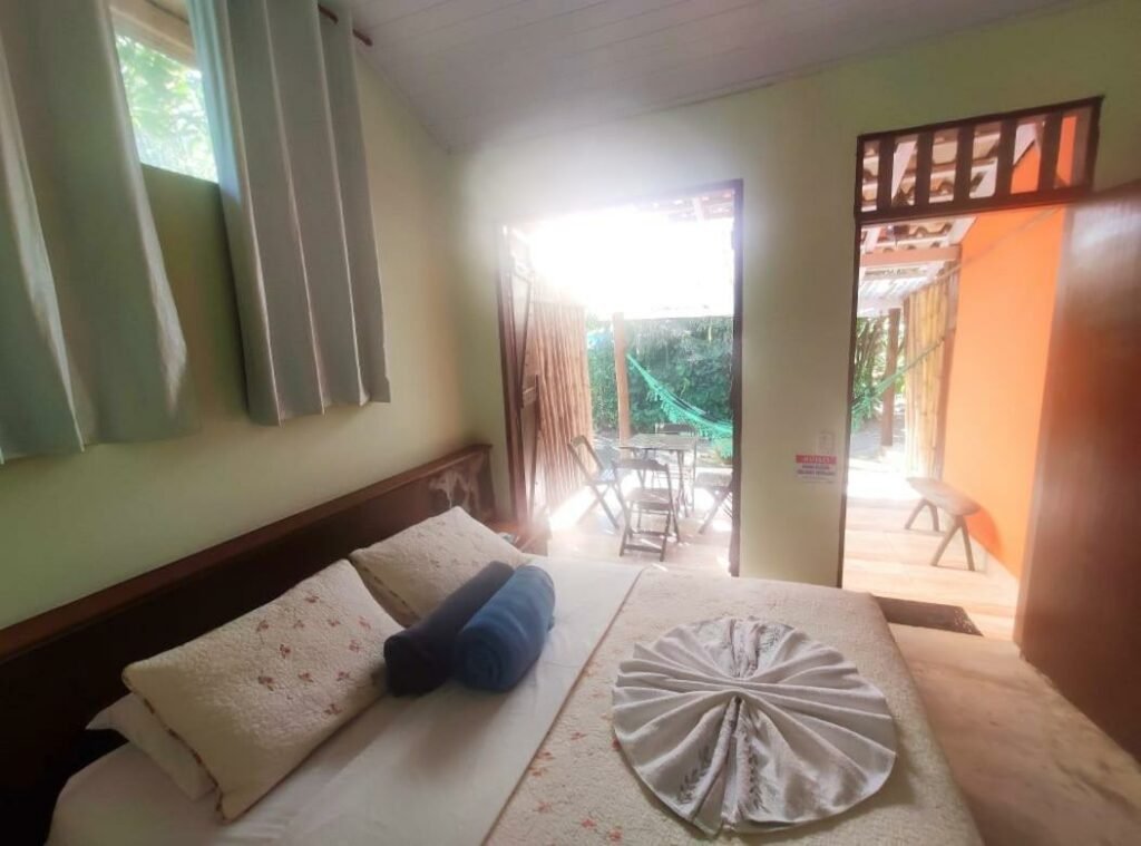 Quarto do Portal do Sabiá Maresias. Uma cama de casal está em primeiro plano, do lado esquerdo. Ao fundo, na varanda térrea, estão uma mesa e uma reede.