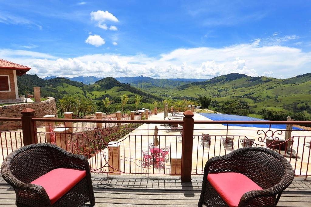 Área externa da Pousada Aldeia Manacás, com uma vista panorâmica de montanhas verdes e céu azul com nuvens brancas. Há também uma piscina com várias espreguiçadeiras em volta e um deck de madeira com duas poltronas em cima