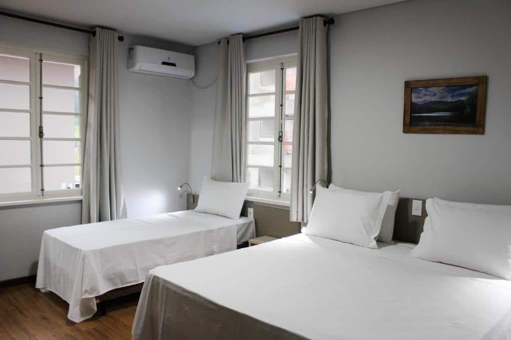 Quarto da Pousada Casarão Schmidt. Uma cama de casal do lado direito, do lado esquerdo uma cama de solteiro, duas janelas e um ar-condicionado. Foto para ilustrar post sobre pousadas pet friedly em Santa Catarina.