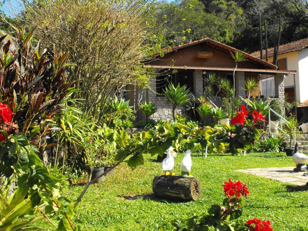 Jardim da pousada cascata dos amores em Teresópolis com plantas e gramado verdes, flores vermelhas e uma casa em tons de cinza e marrom ao fundo durante o dia, ilustrando post pousadas em Teresópolis.