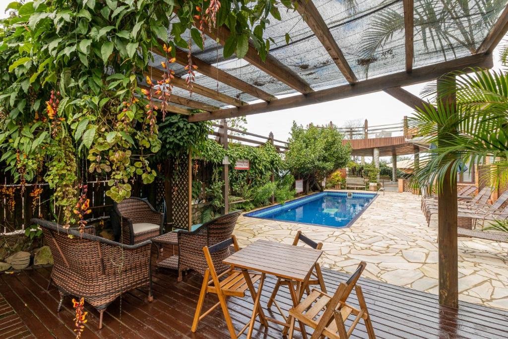Área externa da Pousada Descanso do Rei. Uma piscina no fundo no meio, do lado direito cadeiras de tomar sol. Na parte da frente um deck de madeira com cadeiras e mesas.