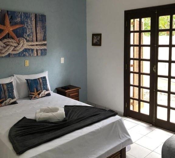 Quarto da Pousada Estrela do Mar. Uma cama de casal está ao lado esquerdo. Ao seu lado estão uma mesinha de cabeceira e a porta para a varanda.