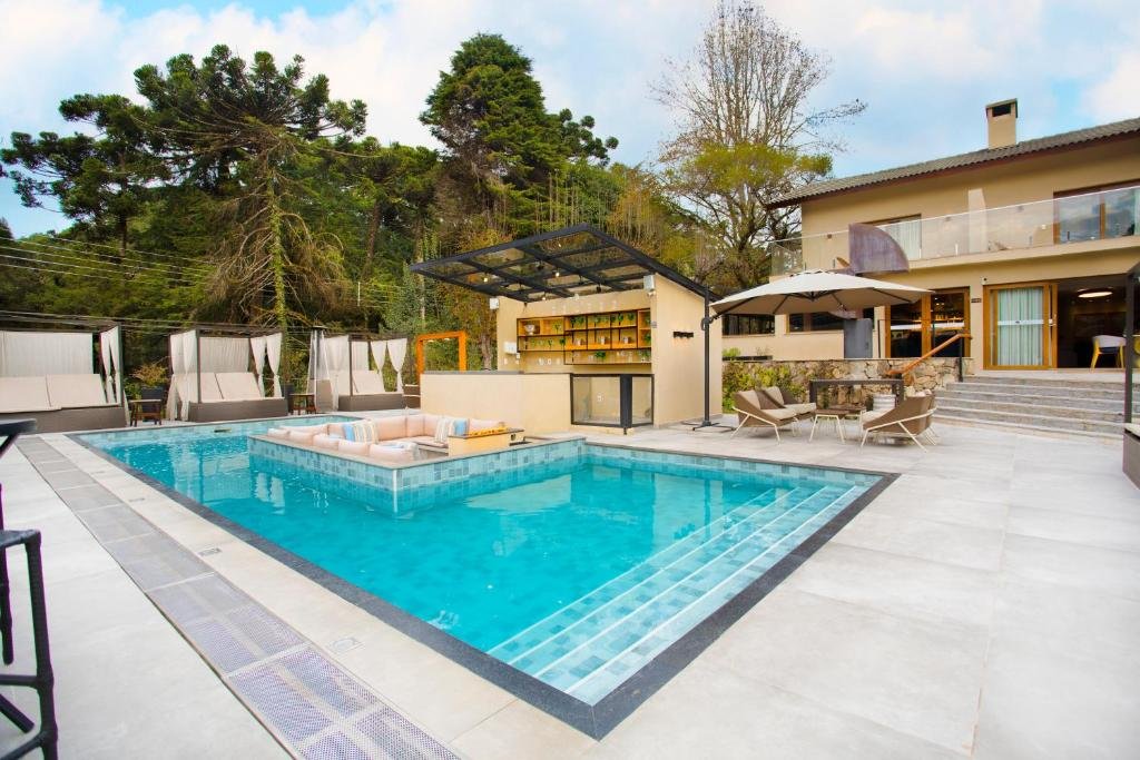 Piscina da Pousada Figueira da Serra cercada por bangalôs com almofadas, há um bar ao lado da piscina, além de algumas cadeiras com guarda-sol, e a propriedade é cercada por árvores