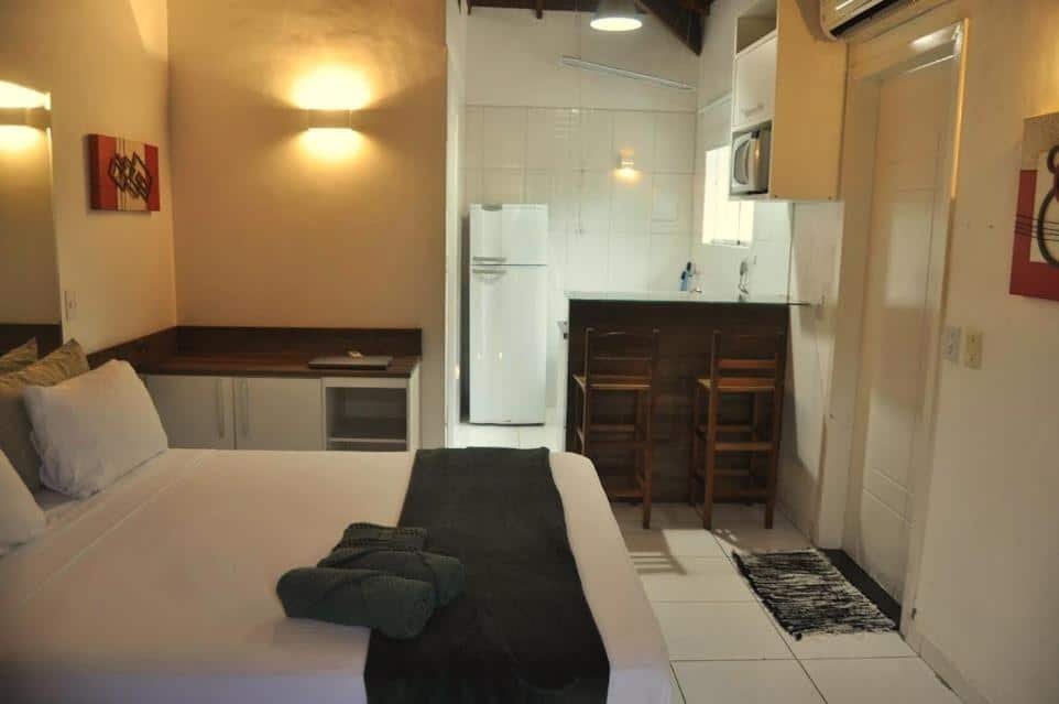 Quarto da Pousada Raio de Sol Maresias. Uma cama de casal está em primeiro plano, ao seu lado um pequeno armário. Ao fundo está uma pequena cozinha e um balcão.