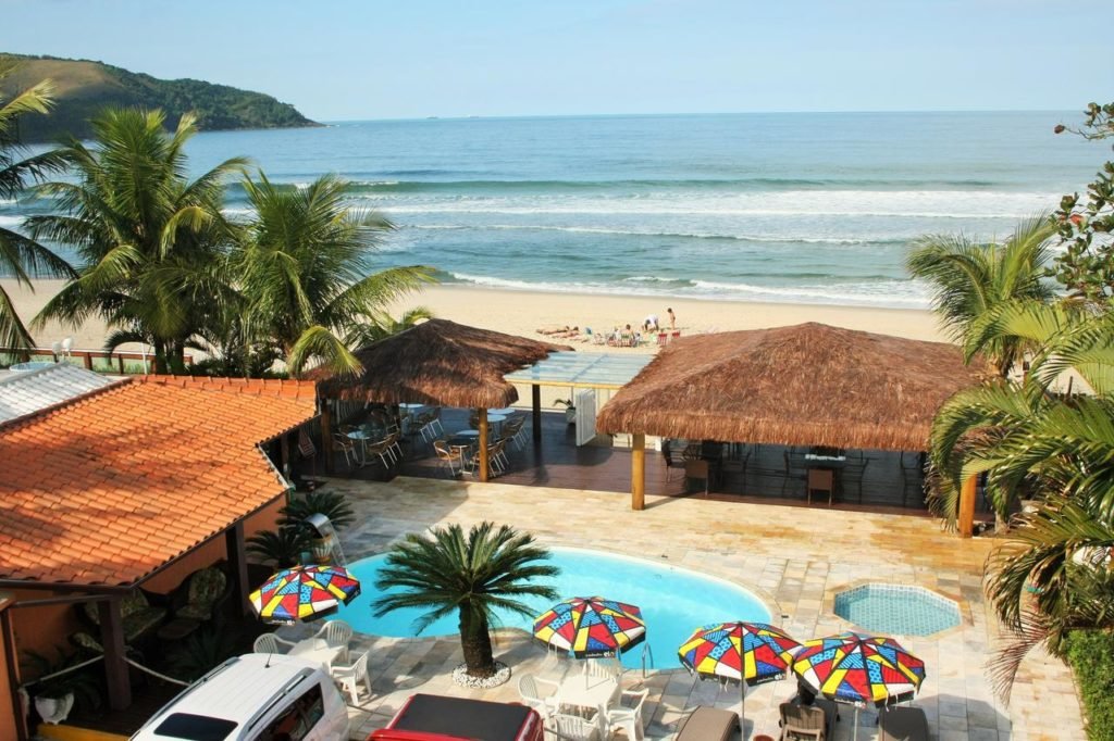 Vista da Praia e piscina na Pousada Beira Mar Maresias. A piscina está rdeada de guarda-sóis, mesas e perto de alguns carros. E frente estão alguns espaços cobertos e logo em seguida a praia.