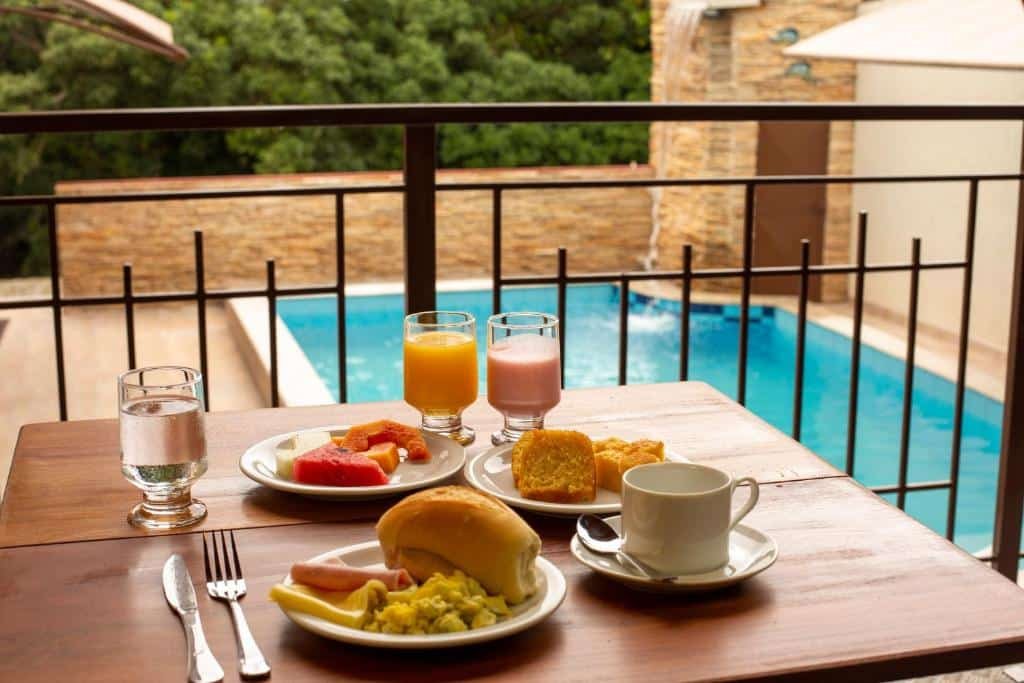 Área de lazer da Pousada nossas Raízes. Uma mesa na frente com pratos de café da manhã, pães, frutas, bolos e frios, com água, café, suco e iogurte. No fundo uma grade da sacada e a piscina.