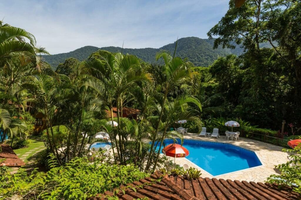 Pisicina da Pousada Pé da Mata Maresias. Ela está no meio de uma área aberta com várias mesas e cadeiras ao redor. Plantas cercam todo o ambiente.