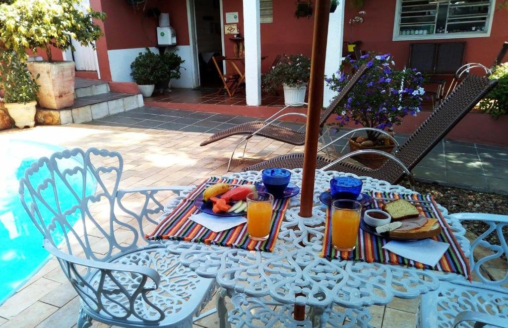 Foto na Piccola Pousada. Na imagem podemos ver uma mesa com guarda-sol na beira da piscina, com pratos de café da manhã em cima que se consistem em uma fatia de bolo, frutas, frios, xícaras de café e suco.