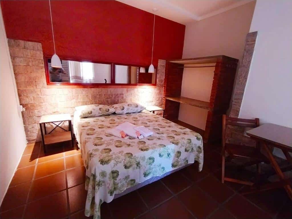 Imagem de um quarto da Piccola Pousada para ilustrar o post sobre pousadas em Águas de São Pedro. Na suíte vemos uma cama box de casal, uma estante de tijolos e madeira do lado direito, e uma pequena mesa de cabeceira. Encostada em uma parede, há uma mesa de madeira com duas cadeiras.