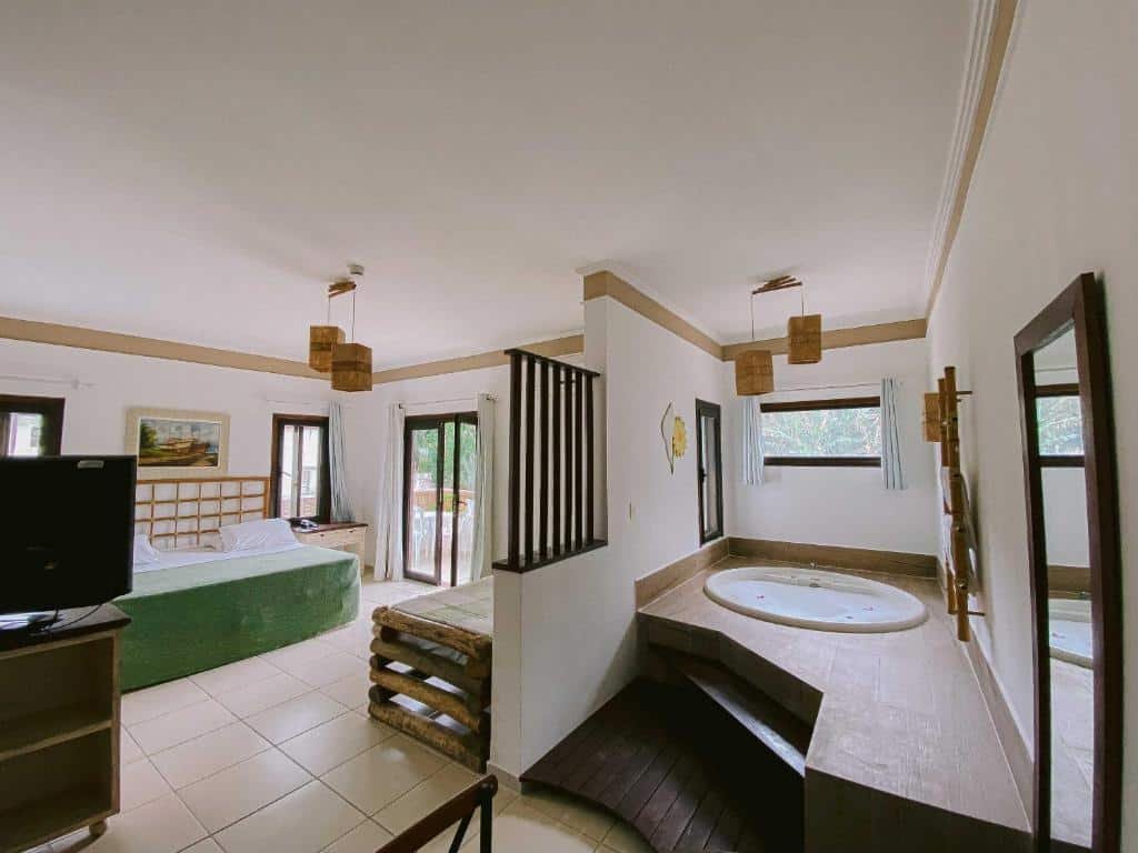 Quarto da Pousada Porto Mare. Ao lado direito está uma banheira redonda de hidormassaem, em uma área elevada. Do lado esquerdo e separados por uma parede que vai até a metade do quarto estão uma cama de casal, uma pequena de solteiro e a televisão. Ao lado da cama de casal há uma porta. Essa é uma das melhores pousadas pet friendly em Maresias.