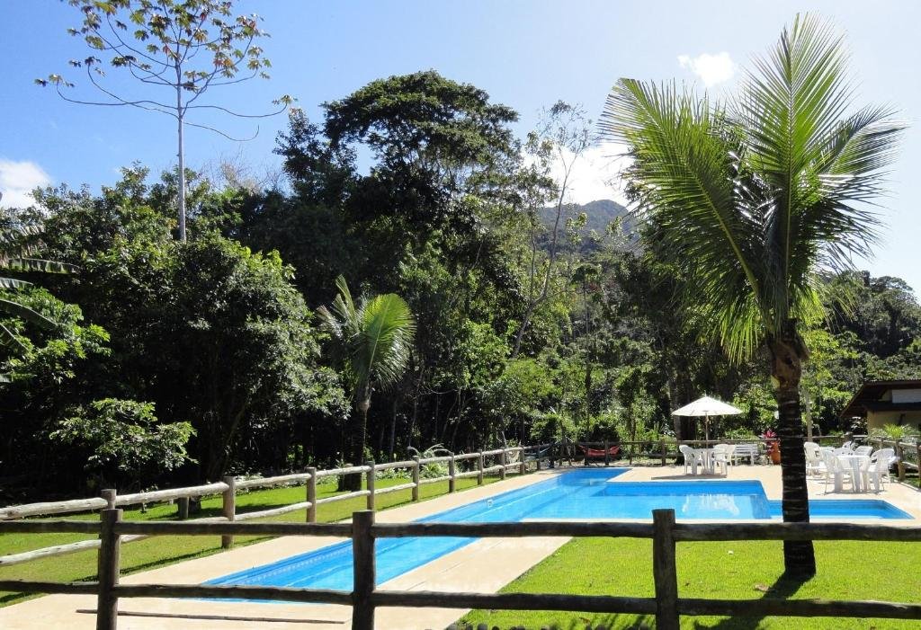 Área externa da Pousada Praia de Itamambuca. Uma piscina no centro com cadeiras e guarda-sóis. Ao redor muita área verde e árvores.